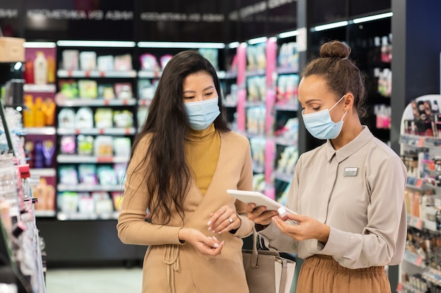 Jovem consultora em máscara protetora rolando em tablet enquanto procura preço de brilho labial procurado por uma das clientes em supermercado de beleza