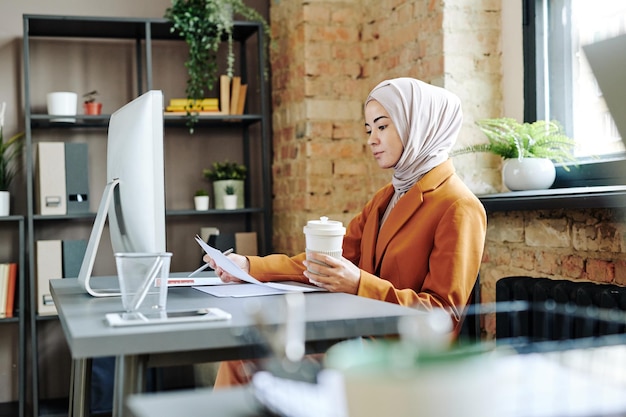 Jovem contadora séria em hijab olhando através de documentos financeiros