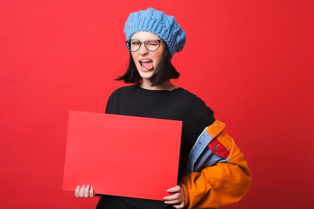 Jovem contemporânea de óculos, mostrando a língua enquanto segura um pôster vermelho vazio