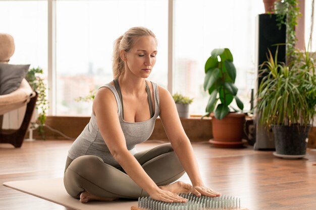 Jovem contemporânea em forma feminina em roupas esportivas, sentada na esteira com as palmas das mãos em almofadas de massagem de ioga durante exercícios de concentração