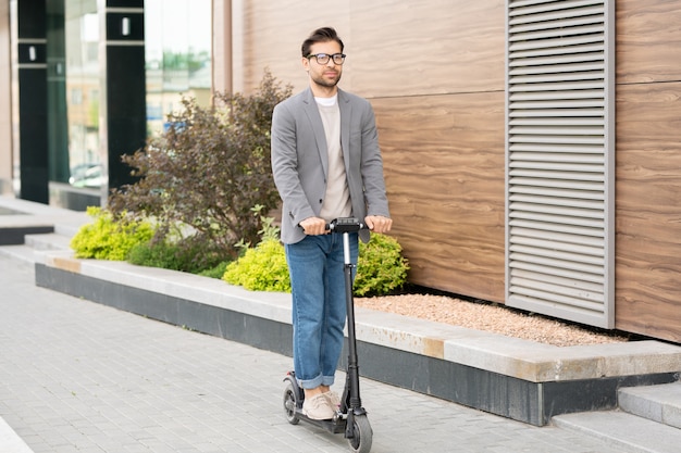 Jovem contemporâneo de jeans, pulôver e jaqueta cinza andando de scooter em ambiente urbano