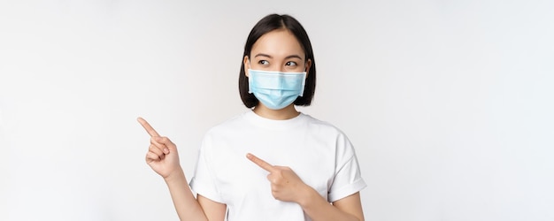 Jovem coreana com máscara facial médica apontando os dedos para a esquerda e olhando para o logotipo mostrando anúncio ou banner sobre fundo branco