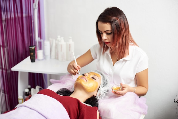 Foto jovem cosmetologista está aplicando a máscara de ouro no rosto de uma cliente morena em um salão de beleza moderno.