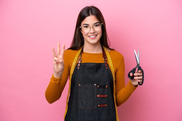 Jovem costureira brasileira isolada em fundo rosa feliz e contando três com os dedos