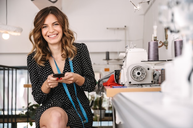 Foto jovem costureira sorridente trabalhando na oficina, usando máquina de costura, usando telefone celular