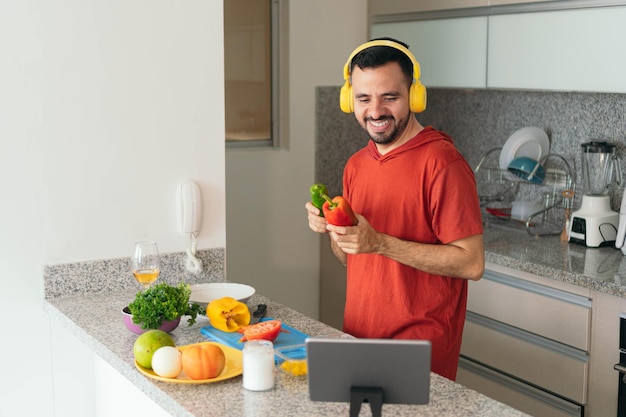 Jovem cozinhando o almoço enquanto faz um bate-papo por vídeo com seu tablet na cozinha moderna