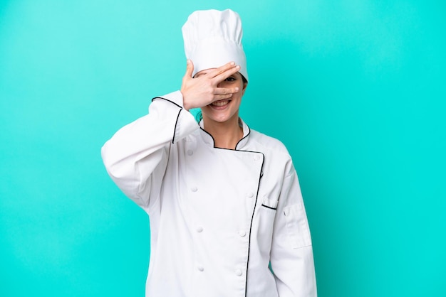 Jovem cozinheira caucasiana isolada em fundo azul cobrindo os olhos com as mãos e sorrindo