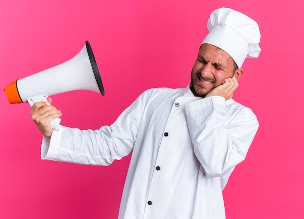 Jovem cozinheiro caucasiano carrancudo com uniforme de chef e boné, mantendo a mão no rosto segurando o alto-falante com os olhos fechados, isolado na parede rosa