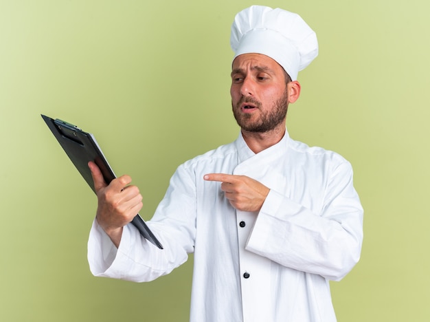 Jovem cozinheiro caucasiano confuso com uniforme de chef e boné segurando e olhando para a prancheta, apontando para ela isolada na parede verde oliva