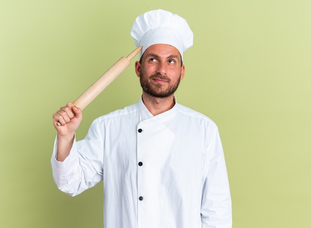 Jovem cozinheiro caucasiano confuso em uniforme de chef e boné tocando a cabeça com o rolo de massa olhando isolado na parede verde oliva