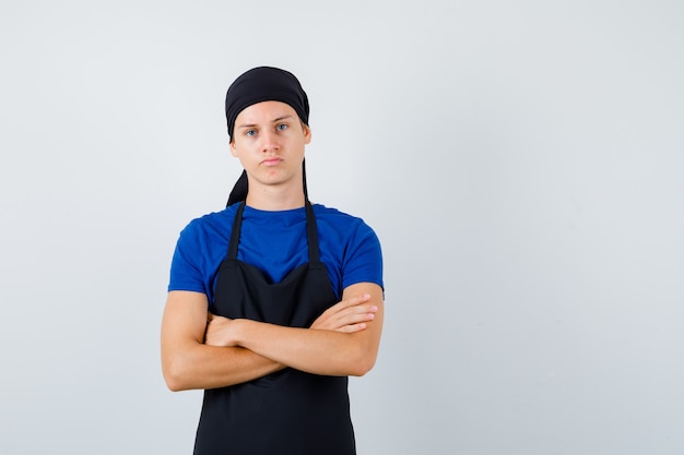 Jovem cozinheiro homem em t-shirt, avental em pé com os braços cruzados e olhando confiante, vista frontal.