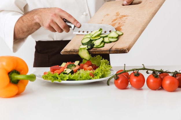 Foto jovem cozinheiro homem uniformizado cortando salada de legumes na placa de madeira isolada na parede branca