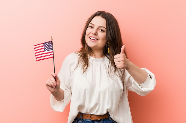 Jovem curvilínea plus size mulher segurando uma bandeira dos Estados Unidos, sorrindo e levantando o polegar