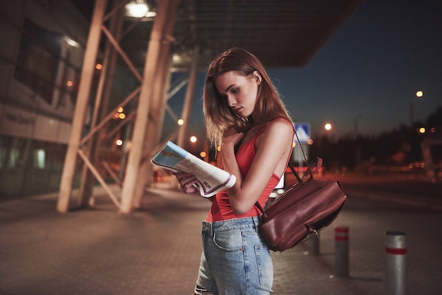 Jovem custa à noite perto do terminal do aeroporto ou estação e lendo o mapa da cidade