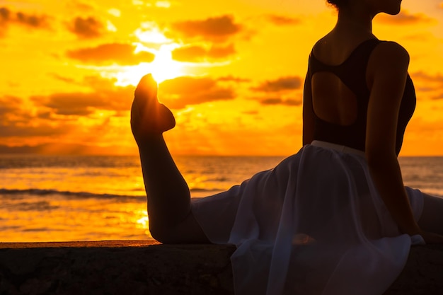 Jovem dançarina ao lado da praia ao pôr do sol fazendo spagata com o mar ao fundo