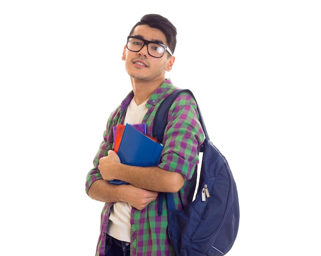 Jovem de aparência inteligente em camiseta branca e camisa quadriculada com mochila azul segurando livros