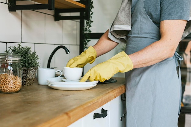 jovem de avental e luvas faz tarefas domésticas lava pratos com toalha no ombro homens trabalho doméstico ajuda doméstica na cozinha elegante em apartamento moderno