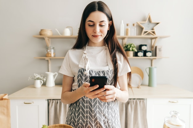 Jovem de avental usando smartphone na cozinha