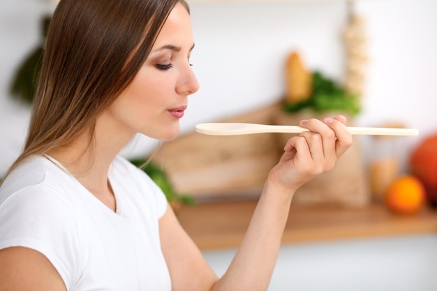 Jovem de avental verde está cozinhando em uma cozinha Dona de casa está provando a sopa com colher de pau