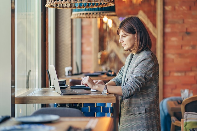 Jovem de blazer com laptop no café perto da janela. Professions é blogueira, freelancer e escritora. Freelancer, trabalhar numa cafetaria. Aprendizagem online.