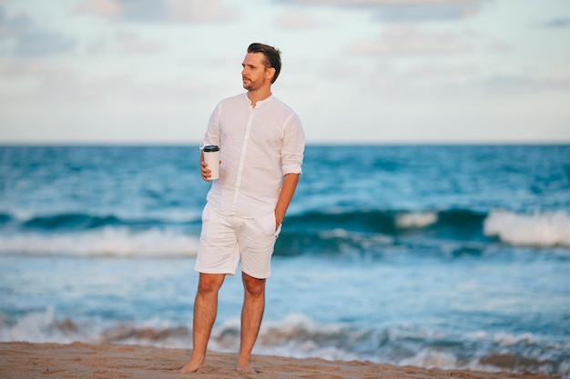 Jovem de branco com café na praia caminhando e aproveitando o entardecer