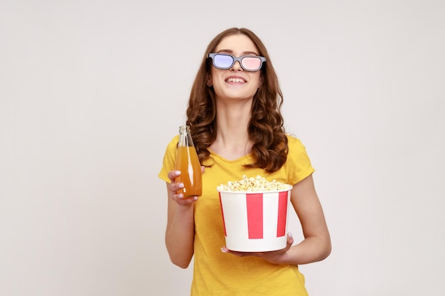 Jovem de cabelos castanhos sorrindo feliz animado adolescente em óculos 3d assistindo filme filme, segure o balde de pipoca e bebendo bebida. tiro de estúdio interior isolado em fundo cinza.