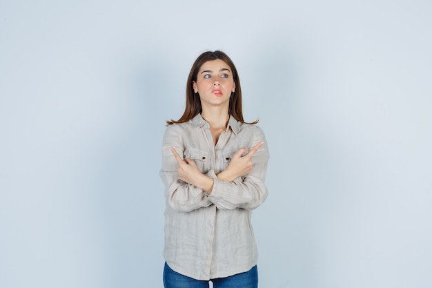 Foto jovem de camisa bege, jeans com dois braços cruzados, apontando direções opostas com o dedo indicador, mordendo os lábios e parecendo indecisa, vista frontal.