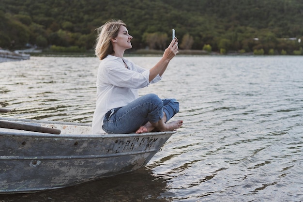 Jovem de camisa branca e jeans com um telefone nas mãos senta-se no barco na margem do lago de montanha