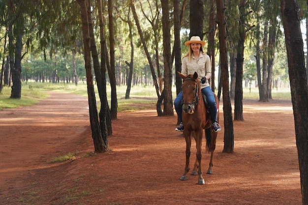 Jovem de camisa e chapéu de palha, cavalgando cavalo marrom no caminho do parque, sorrindo, árvores desfocadas no fundo