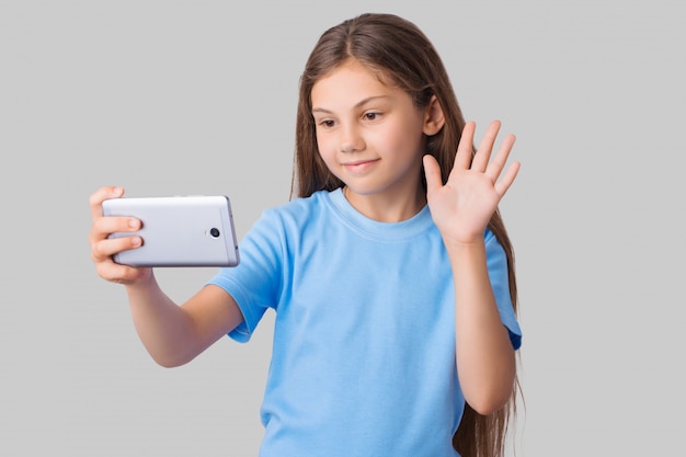 Jovem de camiseta azul, acenando para o telefone móvel, enquanto ela está usando chamadas de vídeo. Pequena estudante com cabelo comprido isolado