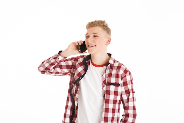 Jovem de camiseta casual usando aparelho segurando o smartphone na mão e tendo uma agradável conversa móvel, isolada sobre parede branca
