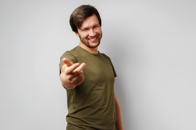 Jovem de camiseta verde sorrindo e gesticulando 'venha aqui' contra o cinza