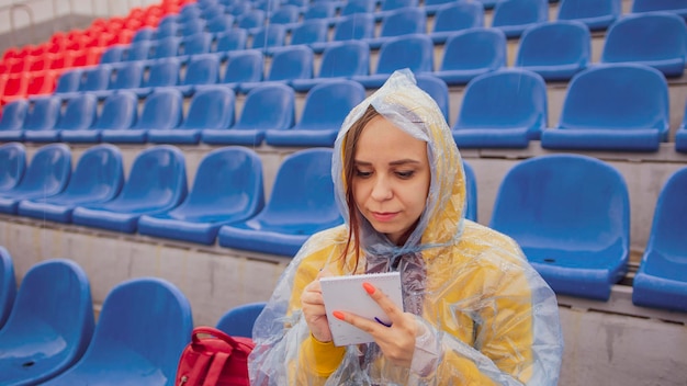 Jovem de capa de chuva com caneta de bloco de notas sentado nas arquibancadas do estádio sozinho em tempo chuvoso Jornalista feminina escrevendo notas durante o treinamento esportivo no estádio de rua