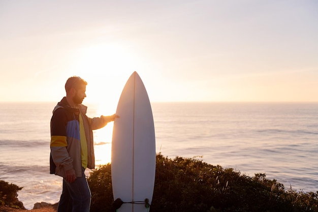 Jovem de casaco segurando sua prancha de surf enquanto olha o pôr do sol à beira-mar lazer e hobbies conceito cópia espaço para texto