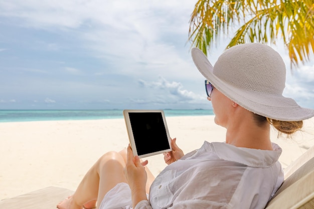 Jovem de chapéu com tablet pc na praia. Escritório em casa praia tranquila praia relaxada trabalho de mulher