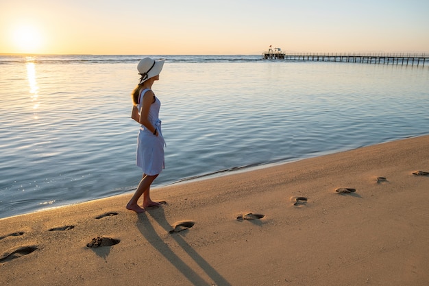 Jovem de chapéu de palha e um vestido andando sozinho na praia de areia vazia na costa do mar por do sol.