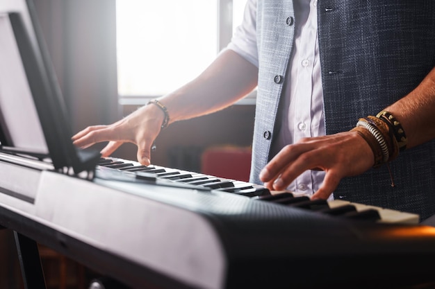 Jovem de colete está tocando piano com as duas mãos em um estúdio de música em casa