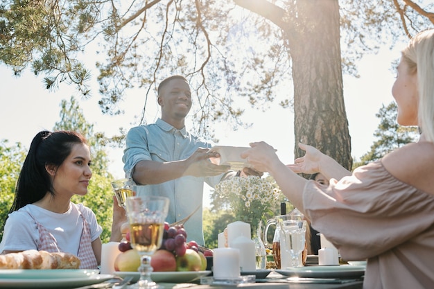 Jovem de etnia africana pegando um recipiente com comida das mãos de sua namorada na mesa servida durante um jantar ao ar livre sob um pinheiro