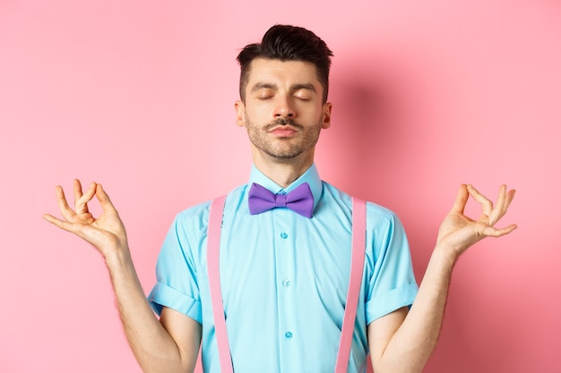 Jovem de gravata borboleta em pé calmo e tranquilo, meditando com as mãos no gesto mudra zen e olhos fechados, pratica ioga para relaxar, em pé sobre rosa.