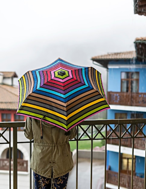 Jovem de jaqueta cáqui por trás com guarda-chuva de arco-íris em pé no terraço na chuva