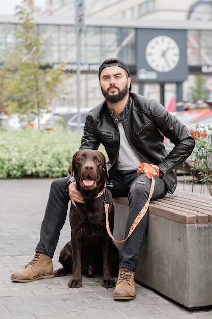 Jovem de jeans preto e jaqueta de couro sentado no banco com um cachorro