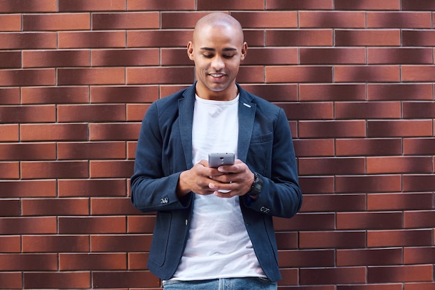 Jovem de lazer em pé encostado na parede navegando na internet no smartphone sorrindo alegre