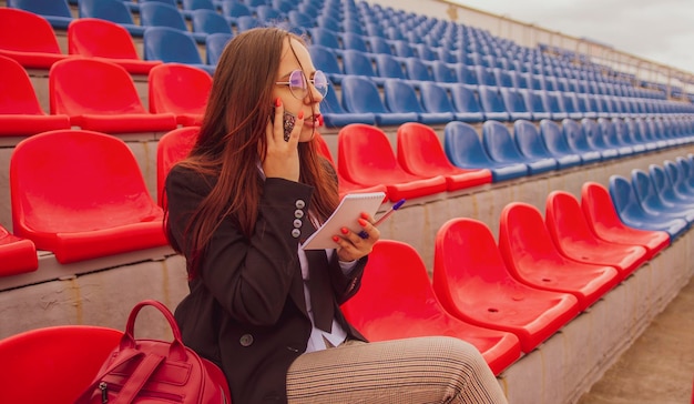 Jovem de óculos com caneta de bloco de notas falando no celular sentado nas arquibancadas do estádio