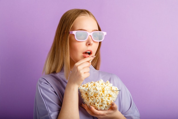 Jovem de óculos de cinema, assistindo filme 3d no cinema. Visualizador de filme de menina adolescente sorridente em copos comendo pipoca isolada sobre uma parede de cor roxa com espaço de cópia.