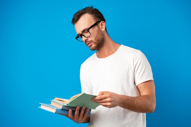 Jovem de óculos, de pé, lendo um livro contra um fundo azul