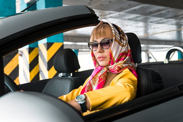 Jovem de óculos escuros, com um lenço na cabeça, dirigindo um carro conversível. loiro modelo de moda coberto subterrâneo estacionamento do shopping center. mulher rica de sucesso elegante elegante em cabriolet.