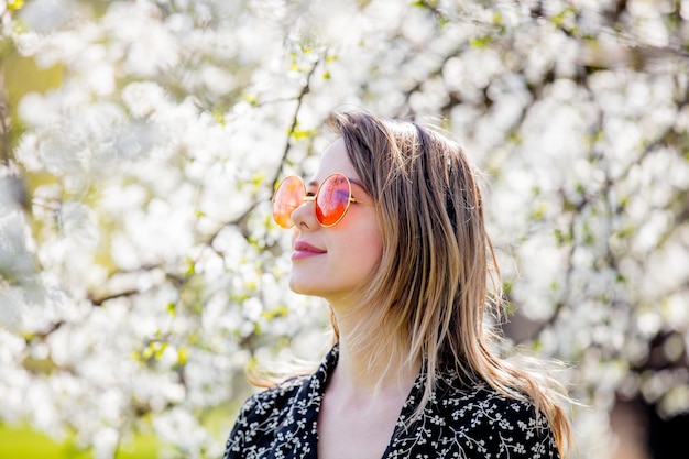 Foto jovem de óculos escuros fica perto de uma árvore florida no parque. temporada de primavera