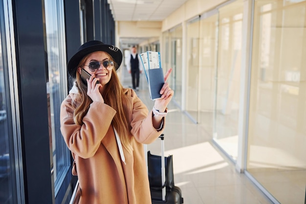 Jovem de óculos escuros, roupas quentes e chapéu em pé dentro de casa no aeroporto com telefone e bilhetes nas mãos.