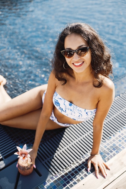 Jovem de óculos escuros sentado perto da piscina e segurando um coquetel. Menina morena vestindo maiô azul e branco
