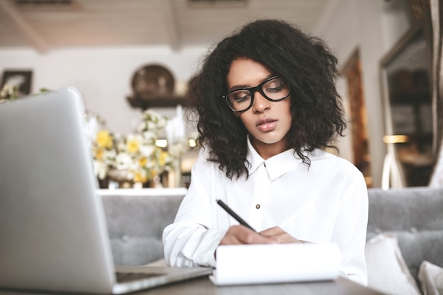 Jovem de óculos, sentada no restaurante e escrevendo pensativamente em seu caderno. Linda garota afro-americana trabalhando em um café com um laptop